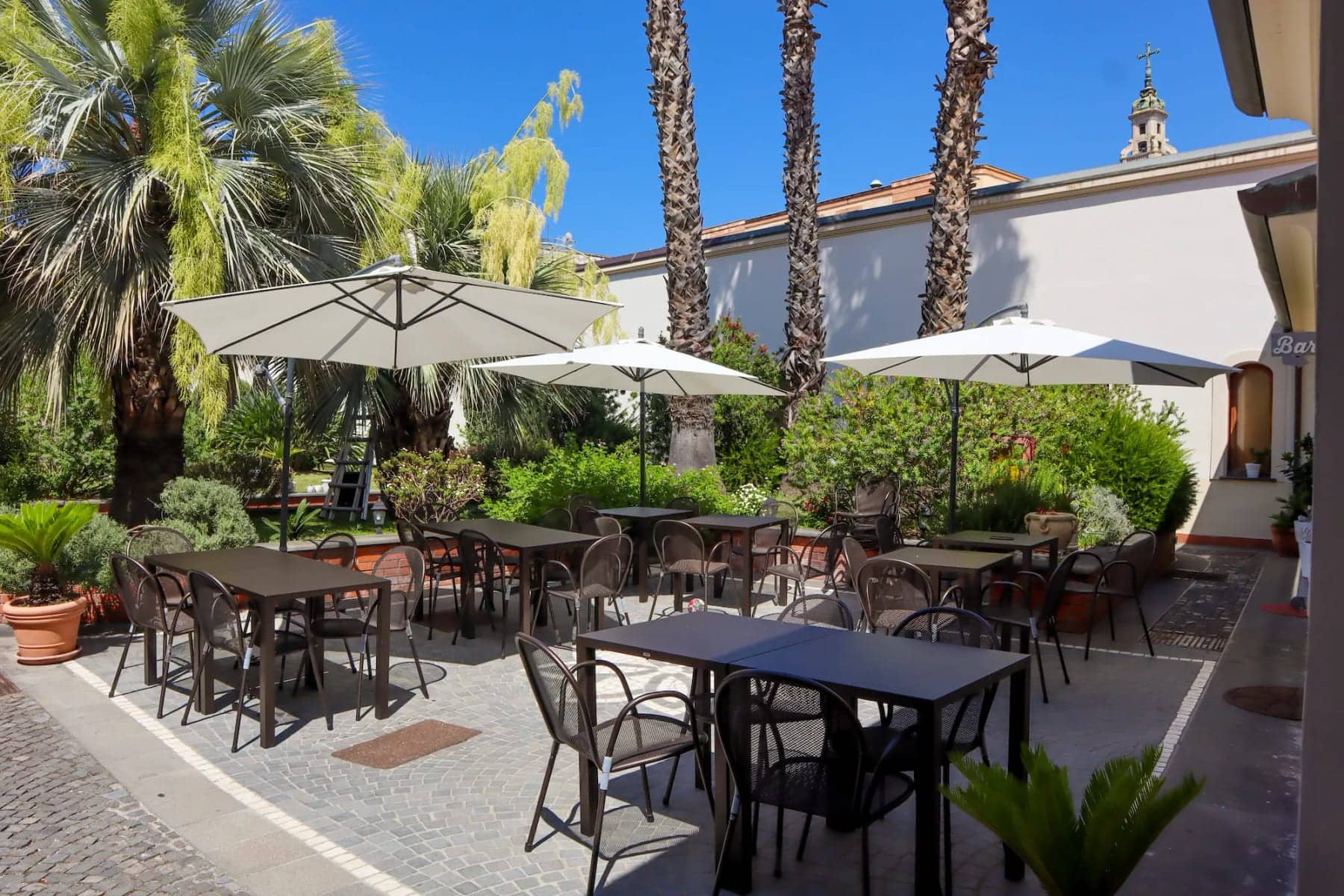 Dehors esterno del Pompei Hostel Deluxe con ombrelloni bianchi, palme e tavoli per la colazione